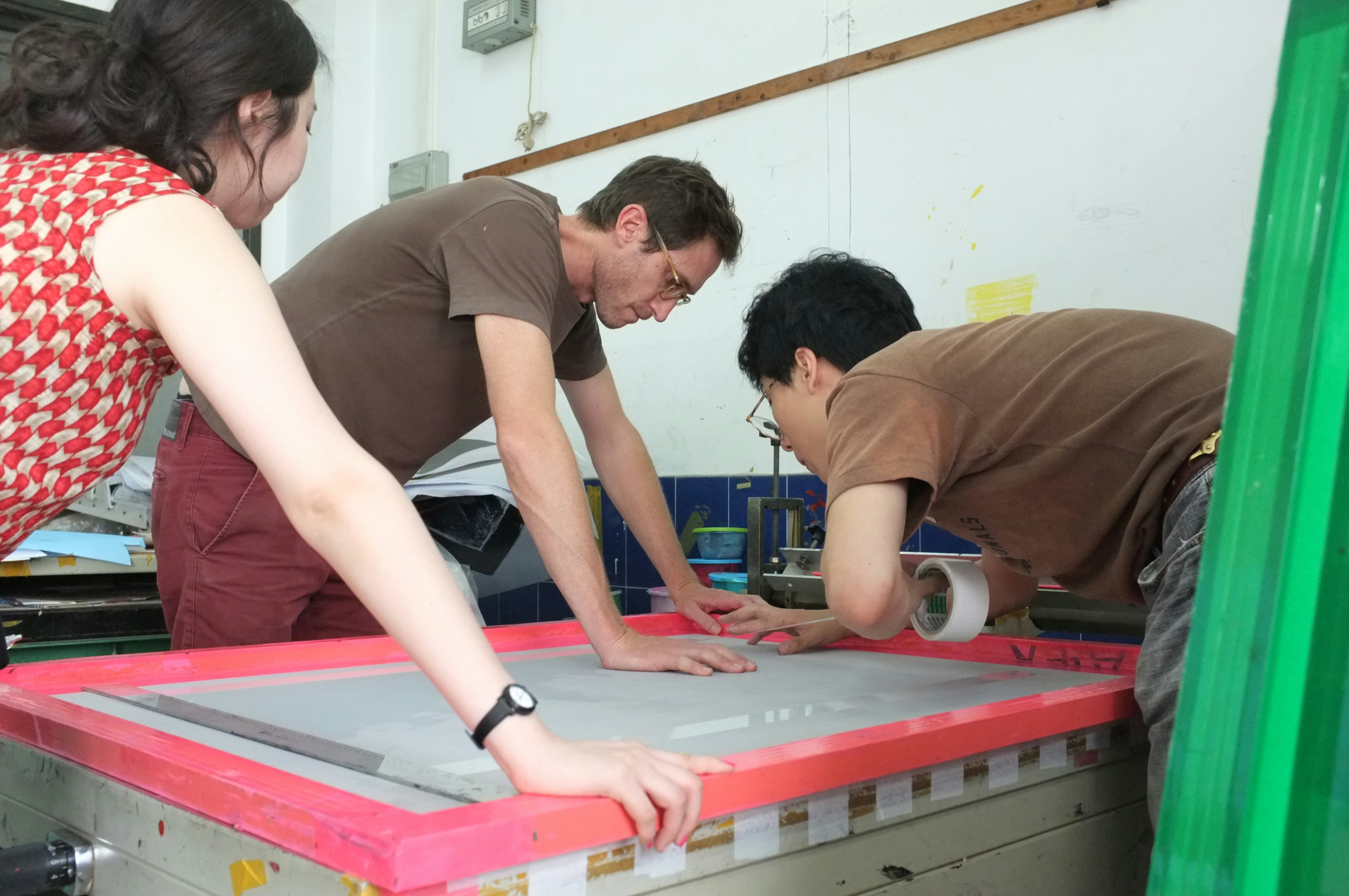 A female-presenting figure and two male-presenting figures leaning over a screen printer.