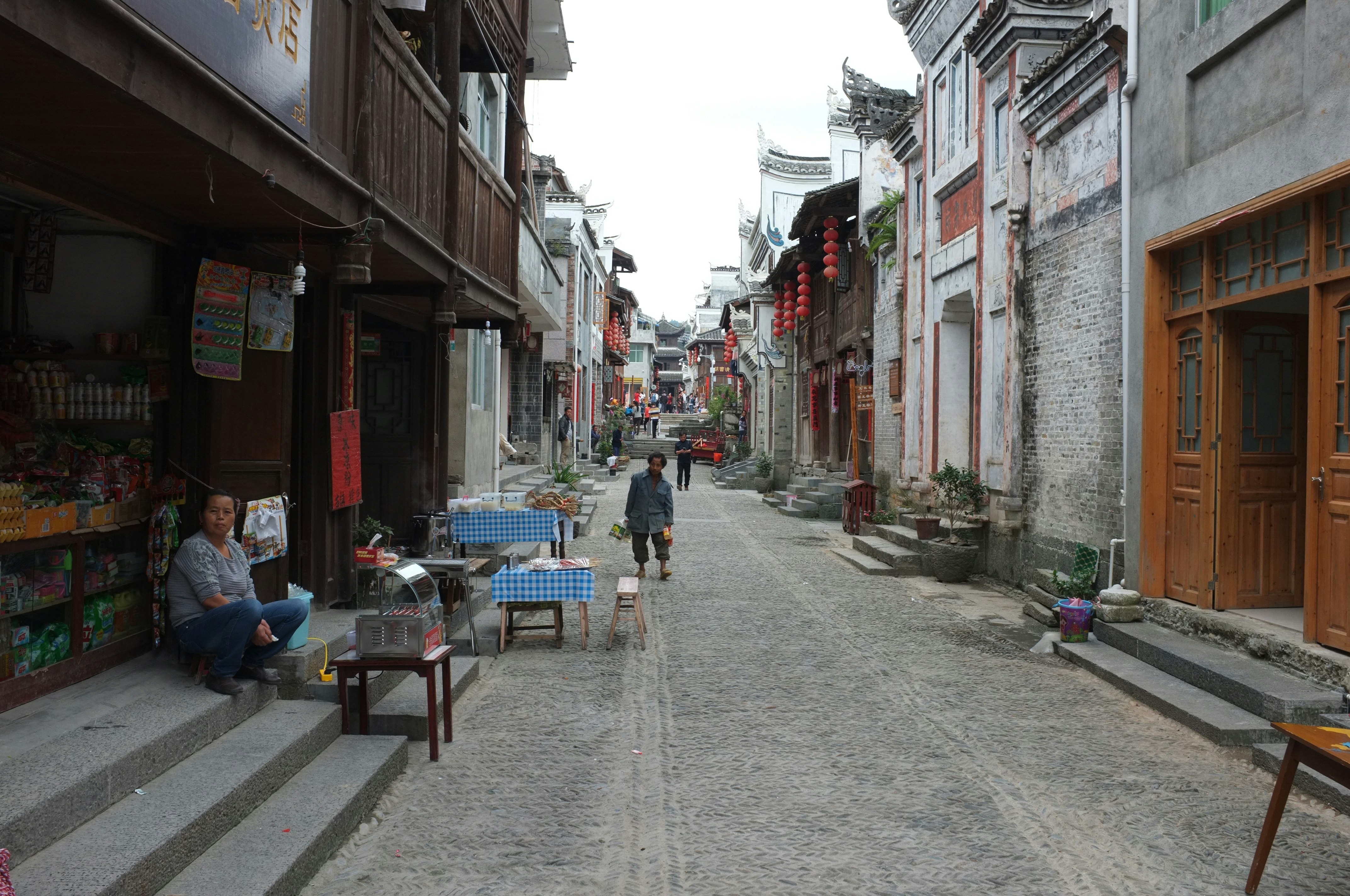 A cobblestone street with residential buildings and shopfronts on each side. A female-presenting figure sits on the steps outside a store.