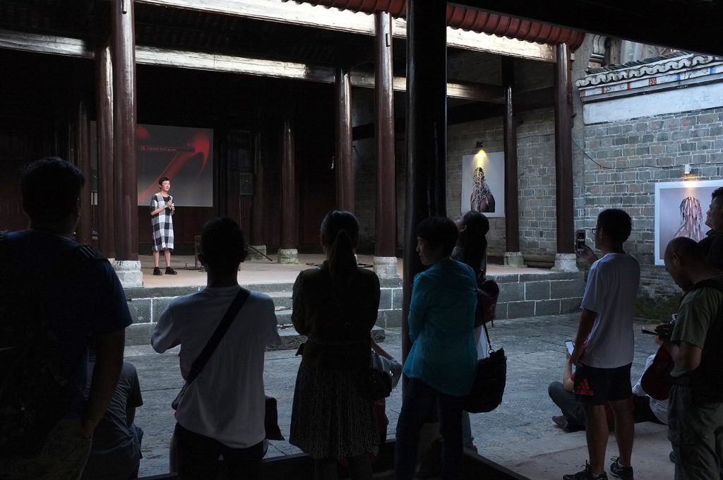 A crowd watches Rainbow, a Chinese female-presenting figure in a black and white checkered dress, standing in a palace.