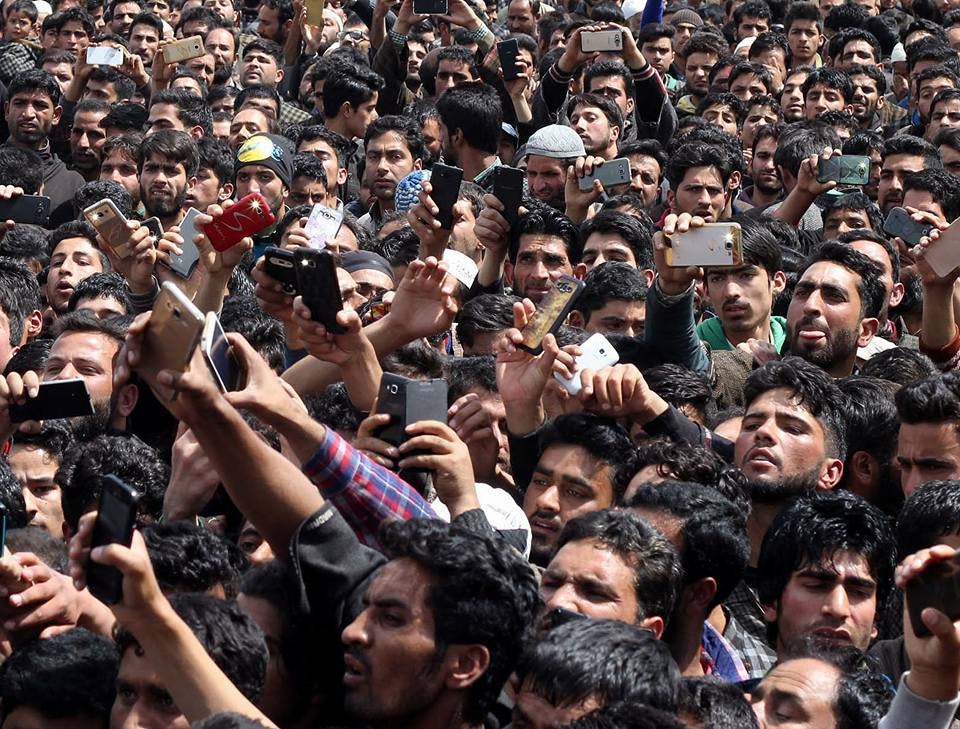 A crowd South Asian male-presenting figures holding up phones to film.