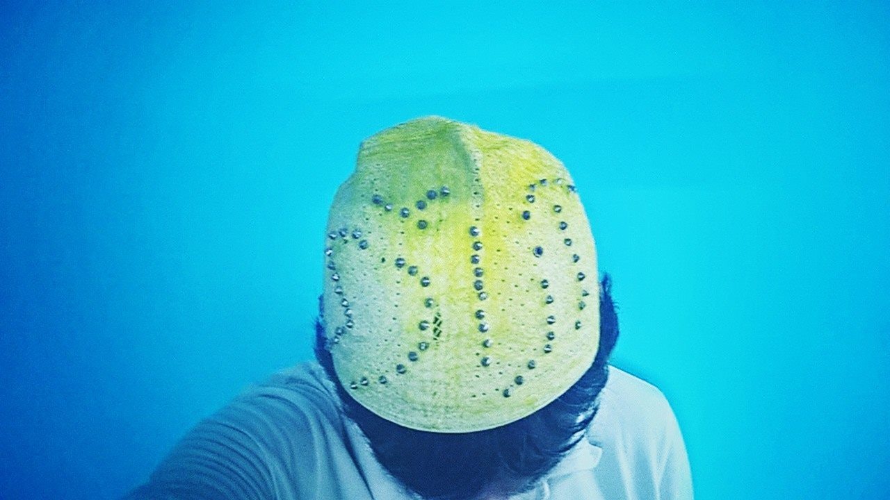 Video still of a male-presenting figure wearing a green hat studded with silver pellets. They stand against a blue background.