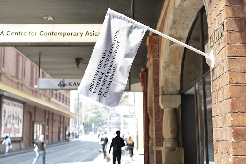 A white flag hanging off a brick wall. The flag reads, 'She sows this Aina with her younger siblings, yet she cannot inherit that same aina.'