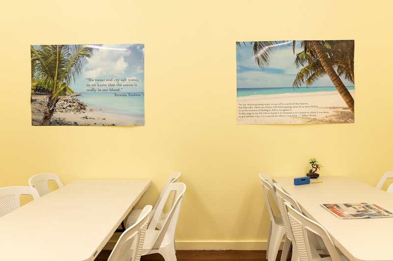 White plastic dining furniture against a yellow painted wall stuck with two posters of palm trees on beaches. One of the posters is printed with a quote from Teresia Teaiwa which reads, 'We sweat and cry salt water, so we know that the ocean is really in our blood.'