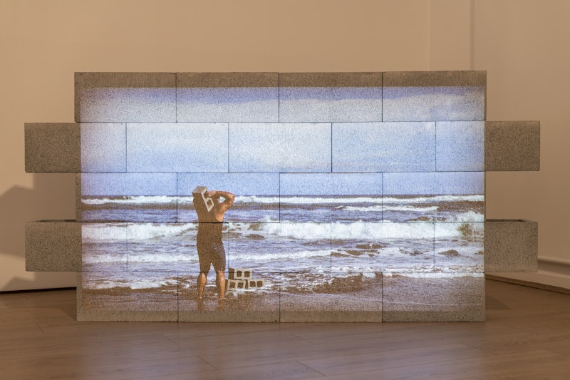 A small cement brick wall projected with a video still of a man holding a concrete block over his shoulders while he faces the ocean. Three concrete blocks are stacked at his feet on the sand.
