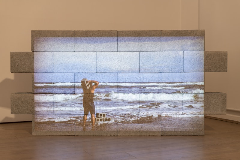 A small cement brick wall projected with a video still of a man holding a concrete block over his shoulders while he faces the ocean. Three concrete blocks are stacked at his feet on the sand.