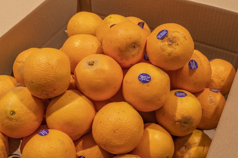 A close-up of oranges with purple stickers. One of the stickers reads, 'Visa Lahi Taha Mahila E 7 $17.70 HE HOUA'