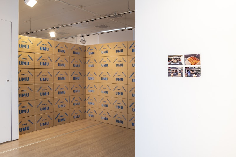 Two walls of stacked cardboard boxes labelled 'UMU' but 'SEASONAL SMALL WORKER' in diagonal blue text above. On the right is a white gallery wall with four printed photos, one of which is an open box of oranges.