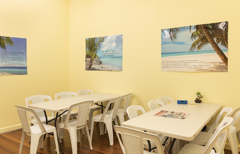 A lunch room with yellow walls, white plastic dining furniture and three posters of palm trees on a beach stuck on the walls.