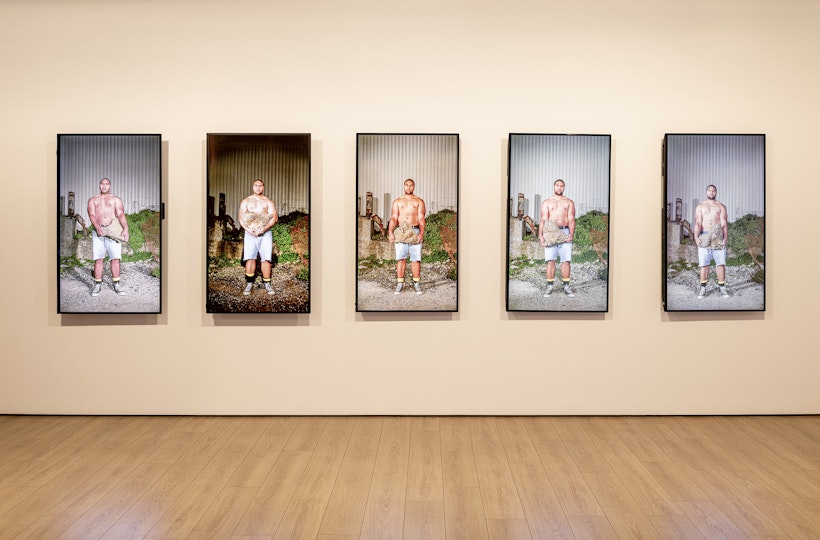 Five television screens on a gallery wall, each showing a shirtless man of Pacific Islander heritage. He is wearing cut-off denim shorts and sneakers, while carrying a large rock against an industrial backdrop