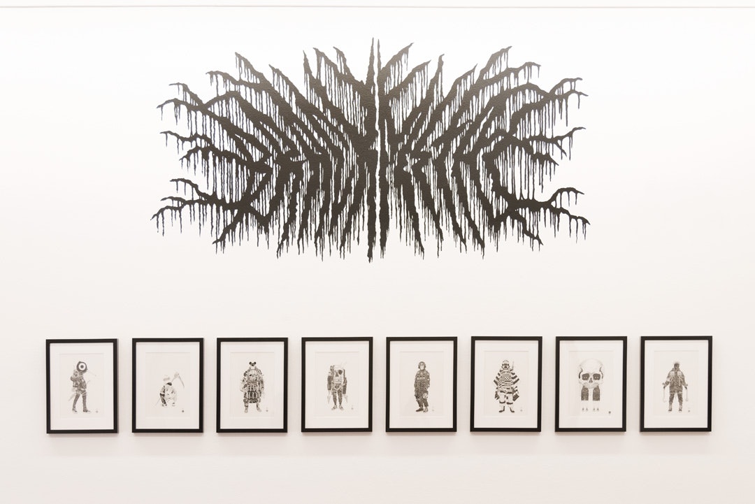 Eight framed black ink drawings on a white gallery wall. Above these drawings is a mural composed of black lines resembling tree branches or veins, stretching from a spine outwards.