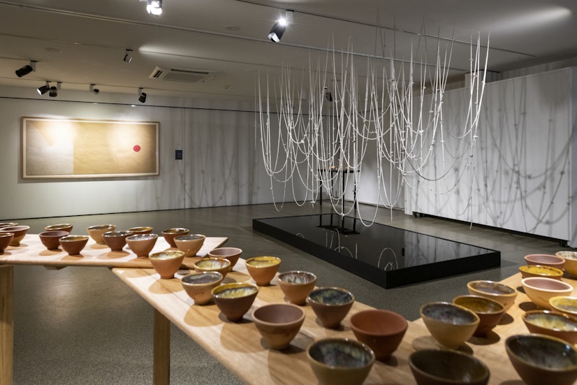 Black exhibition signage that reads, 'Drawn by stones: Dean Cross, Ray Chan See Kwong with Chuen Lung Community Members, Penny Evans, Ruth Ju-Shih Li, Wen-Hsi Harman with Lakaw, Dogin, Palos, Lisin, and Byimu, Jody Rallah.' Behind are transparent plinths and a wall painting in brown ochre. 