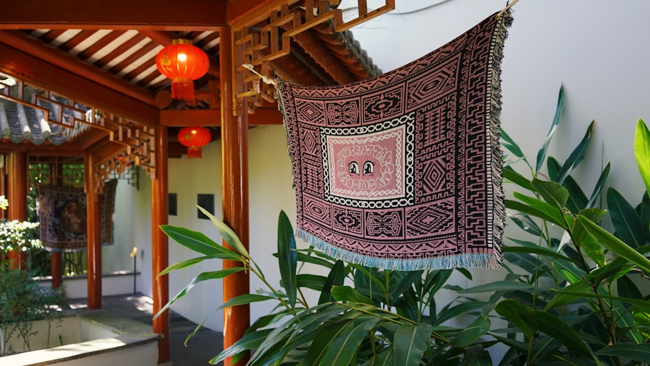 A woven tapestry of a pair of cartoon eyes surrounded by red and black patterning, hanging above some plants in a garden