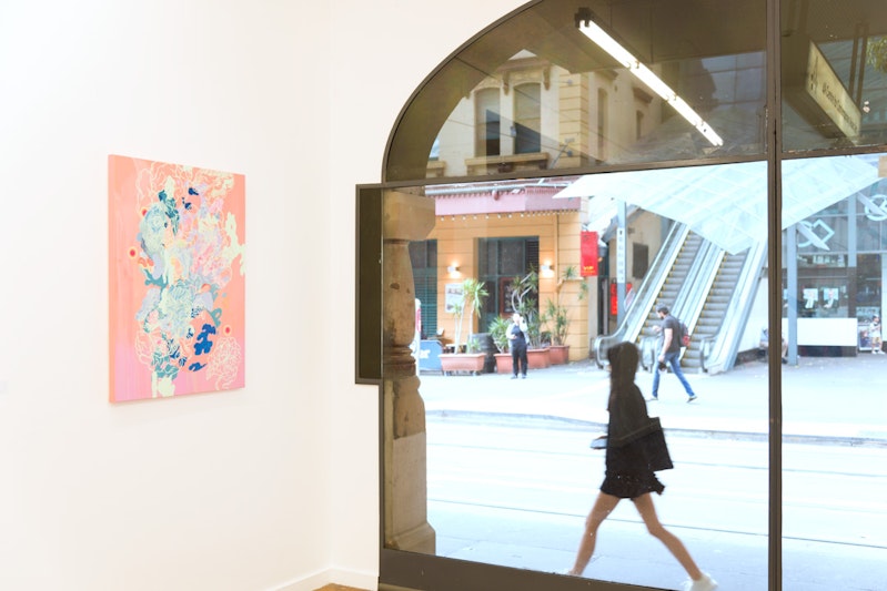 A painting with a peach-coloured background and pastel green, teal and deep blue clouds painted over, hung on a white gallery wall next to a glass window. A girl with long black hair walks past the glass window.