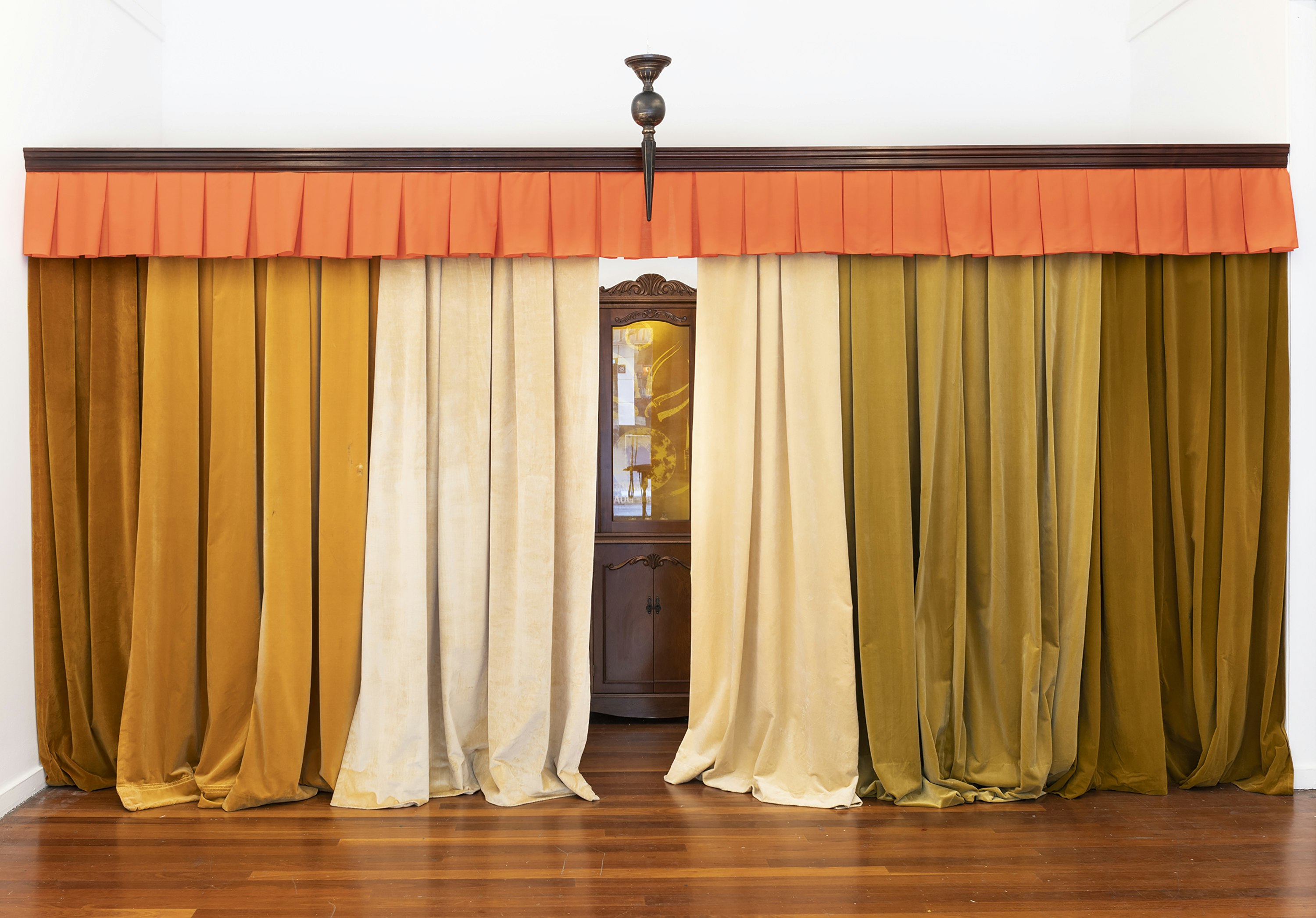 A curtain sewn from beige, light orange and brown fabrics under a bright orange curtain header. The curtain sweeps a hardwood timber floor. Through a middle parting in the curtain is a wooden glass cabinet.