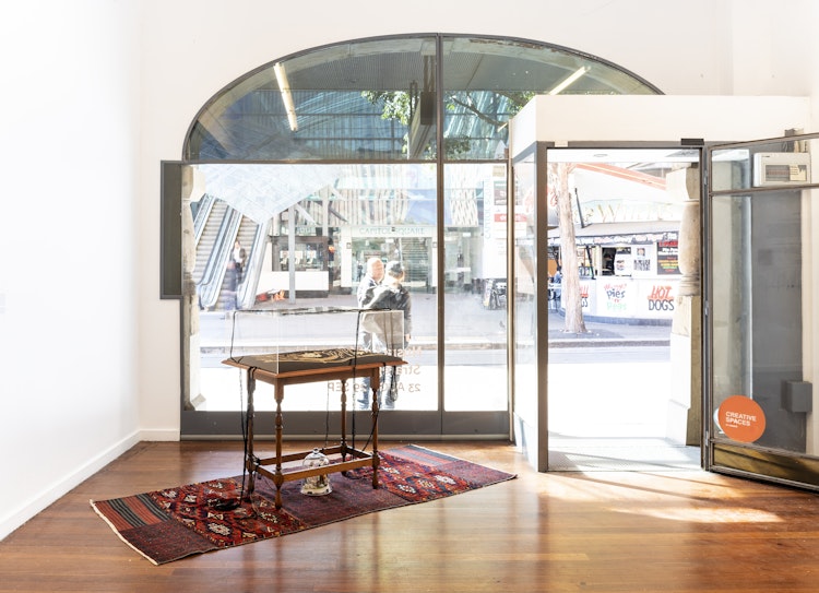 A wooden table covered in glass stands on a red embroidered velvet mat in a light-flooded white gallery space, with a glass door that opens out onto the street outside. Under the table is a glass cloche