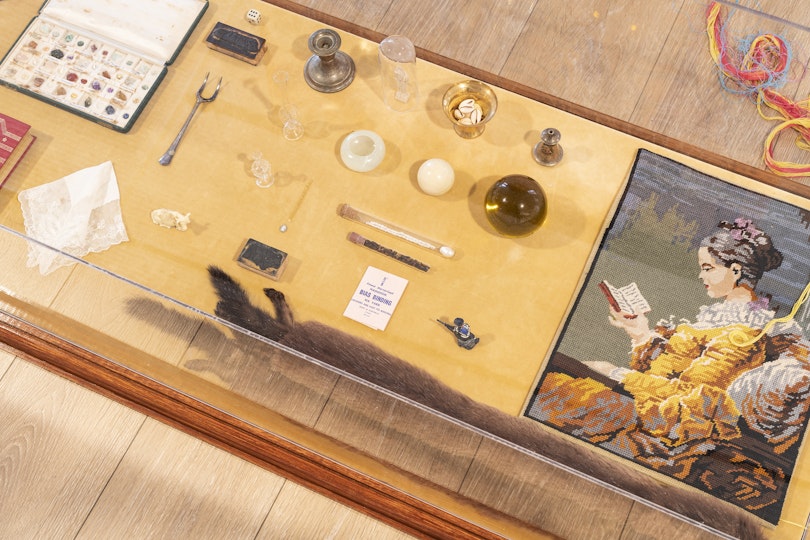 A glass cabinet displaying small objects, including a gilded bible, a lace handkerchief and an ivory orb. A stuffed animal resembling a ferret lies prone next to an embroidered image of a regal woman in colonial dress reading from a small book