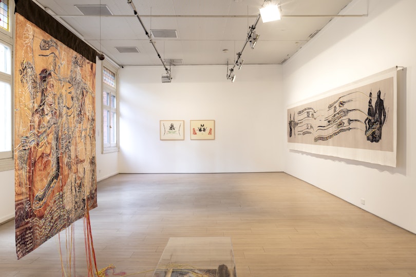 A white gallery space with an orange beaded tapestry, two framed lithographs against a back wall and a long digital print on fabric stretched across the right wall