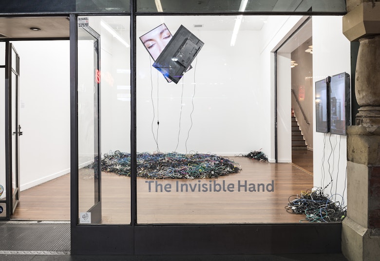 A gallery glass front with the decal that reads 'The Invisible Hand' looks into a space with a mound of electrical cords on the floor and two hanging television screens from the ceiling above. A face with closed eyes is displayed on one of the screens while the other screen is overlapped on the face's mouth as both screens are kissing