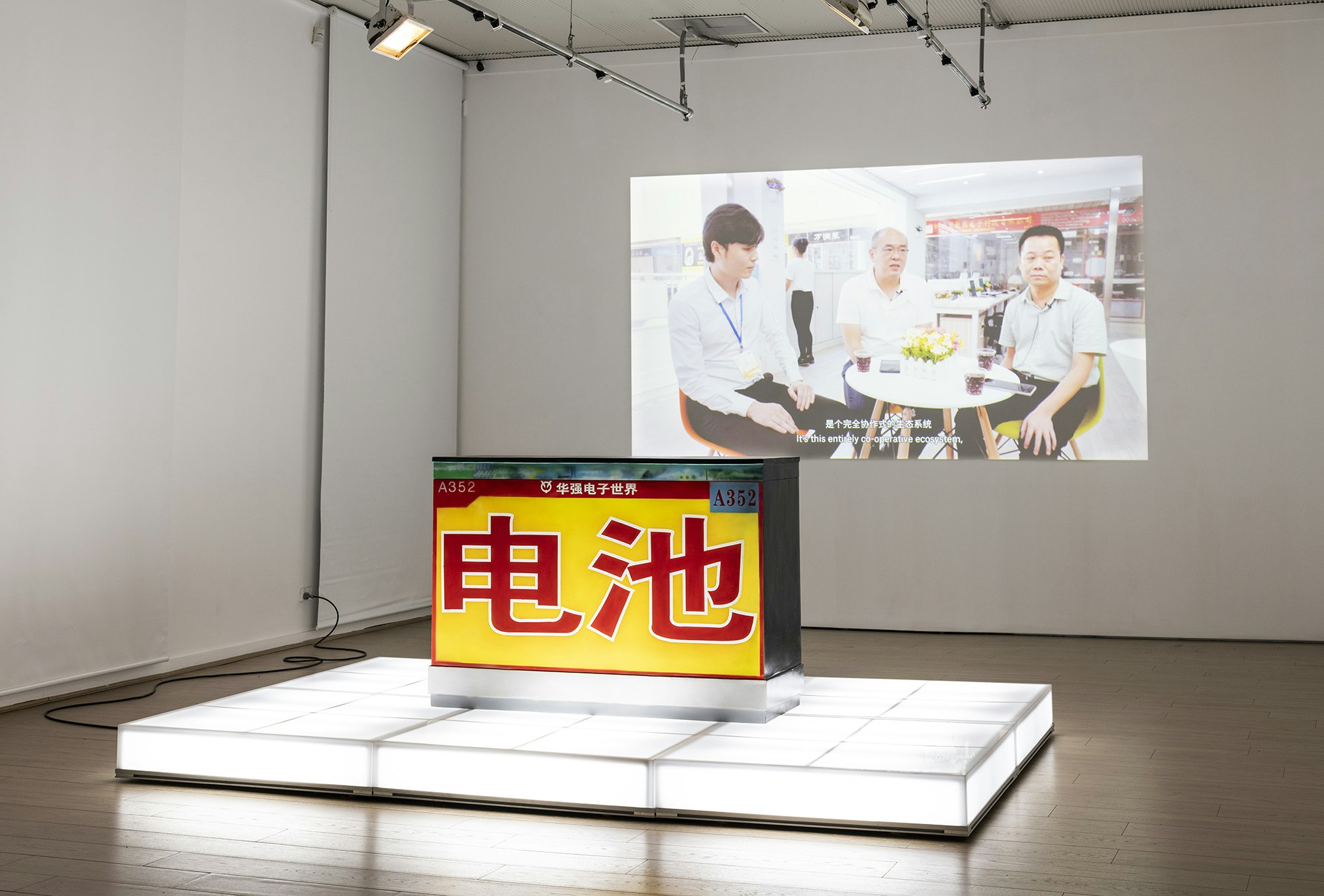 A yellow sculptural block emblazoned with red Chinese characters stands on an illuminated white plinth, with a video still of three seated male-presenting figures projected onto the wall behind