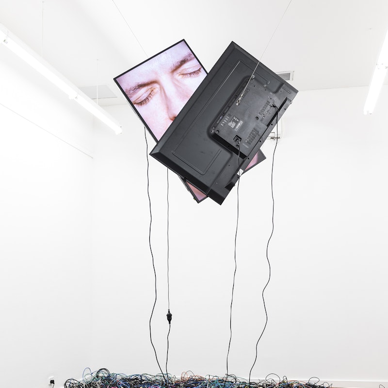 Two television monitors are suspended from a gallery ceiling, one broadcasting a kissing face overlapped with the other television monitor. Cords from the monitors hang down into a pile of electrical cords on the gallery floor.