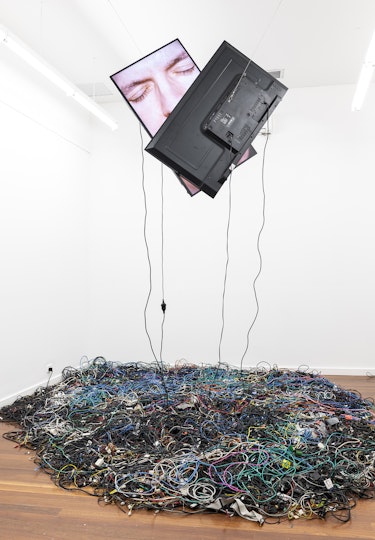 Two television monitors are suspended from a gallery ceiling, one broadcasting a kissing face overlapped with the other television monitor. Cords from the monitors hang down into a pile of electrical cords on the gallery floor.