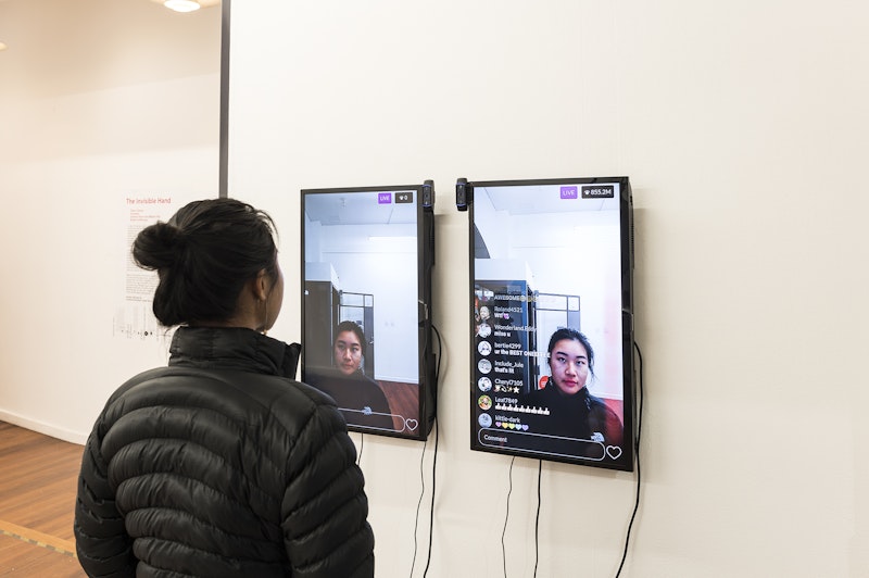 A femme-presenting figure in a black down jacket and her hair in a bun looks into a webcam broadcasting an Instagram livestream of her face onto two television screens mounted on a white gallery wall