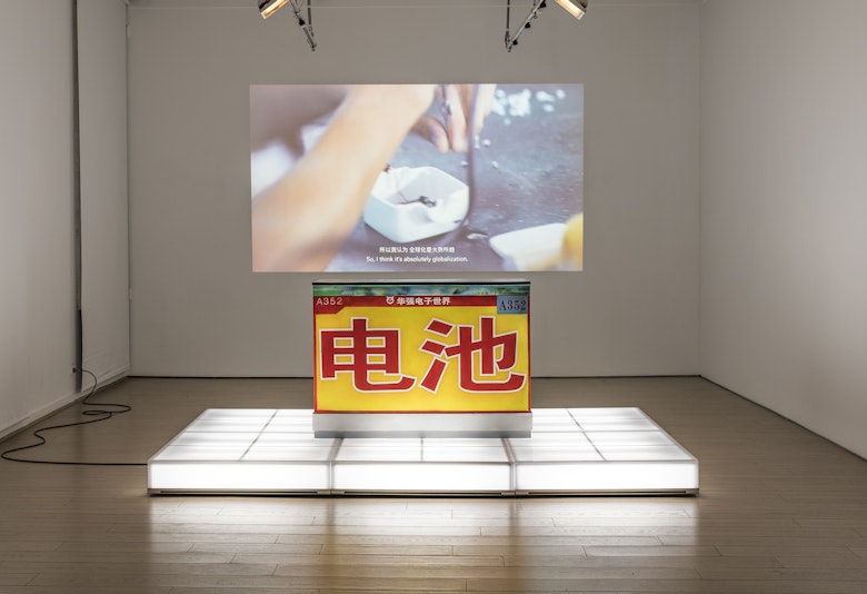 A yellow sculptural block emblazoned with red Chinese characters stands on an illuminated white plinth, with a video still of hands projected onto the wall behind.