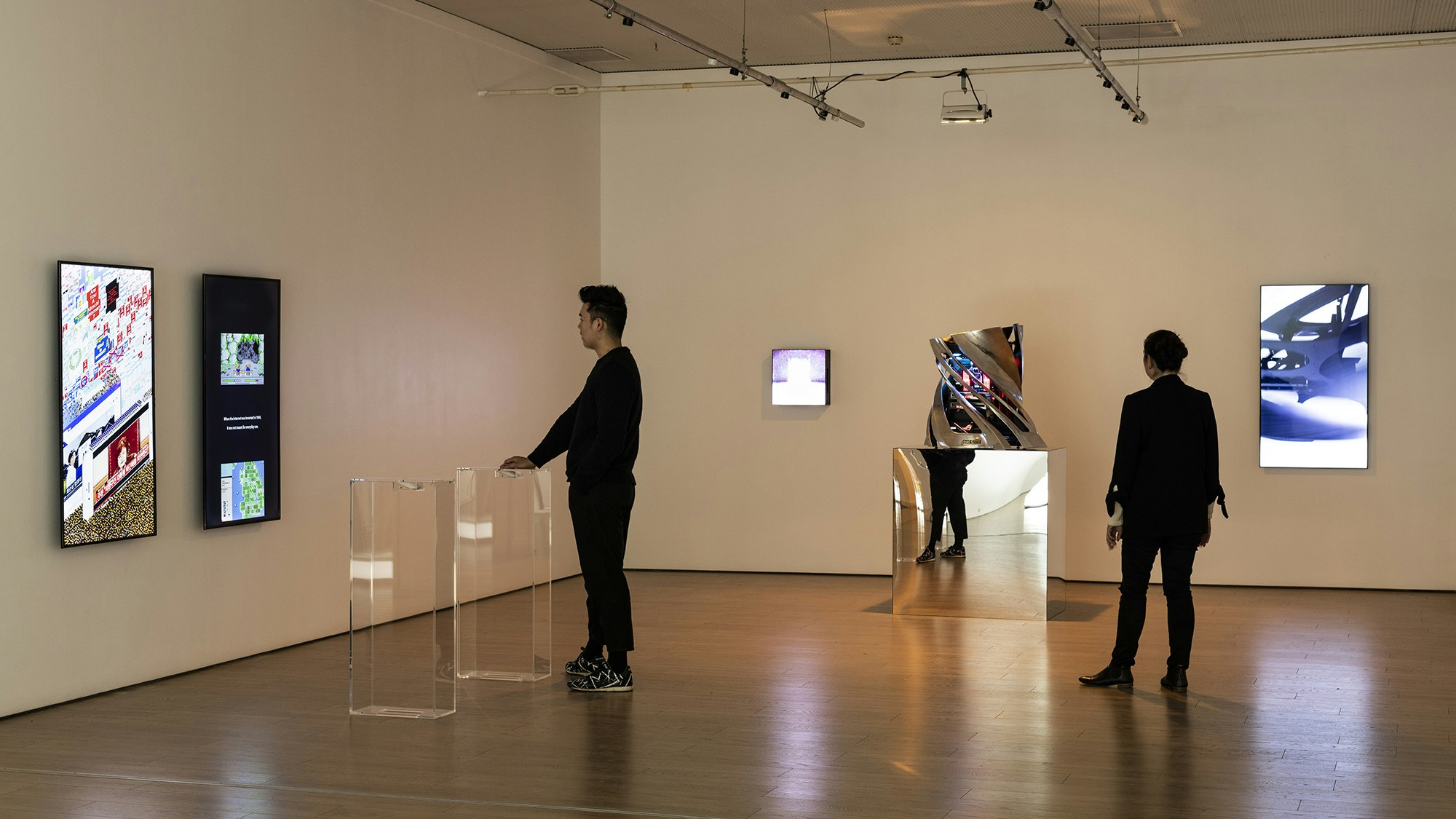 Two figures in black stand in an art gallery. One has his hand on a clear perspex plinth where a large scroll wheel is embedded. He looks at the two vertical LED screens in front of him. The other figure looks at a twisted platinum sculpture on a reflective plinth.