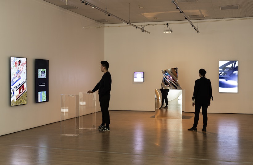 Two figures in black stand in an art gallery. One has his hand on a clear perspex plinth where a large scroll wheel is embedded. He looks at the two vertical LED screens in front of him. The other figure looks at a twisted platinum sculpture on a reflective plinth.
