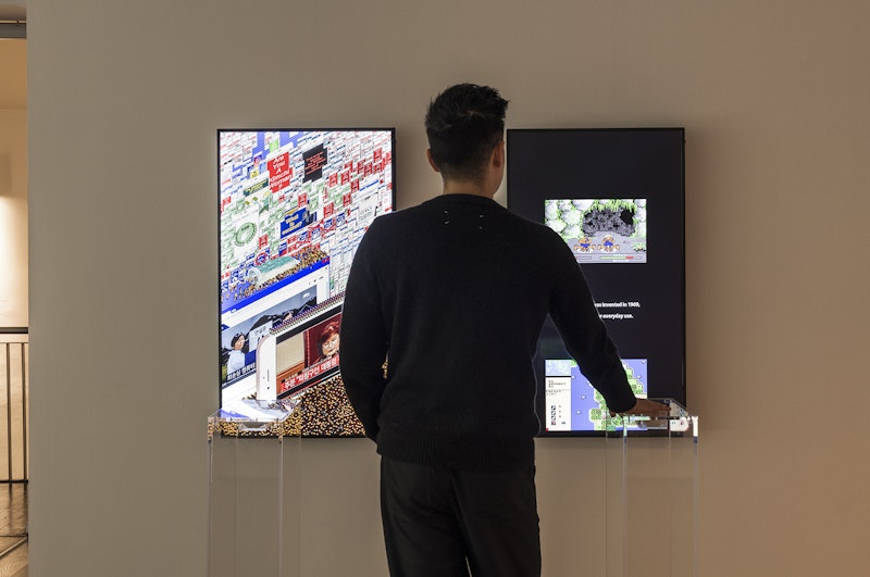 A male-presenting figure in black looks at two LED screens in front of him, with his right hand on a large scroll wheel embedded into a clear perspex plinth. The left LED screen depicts an 8-bit computer game graphics scene