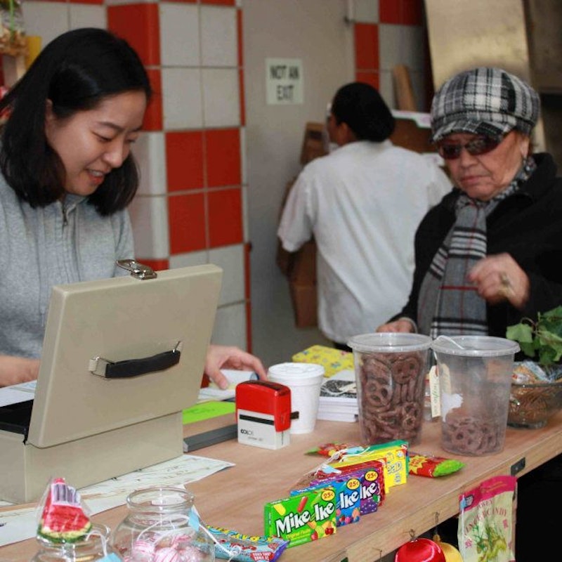 Minja, a South Korean femme-presenting figure in a grey hoodie, stands over a kitchen bench decorated with kitchen ingredients.