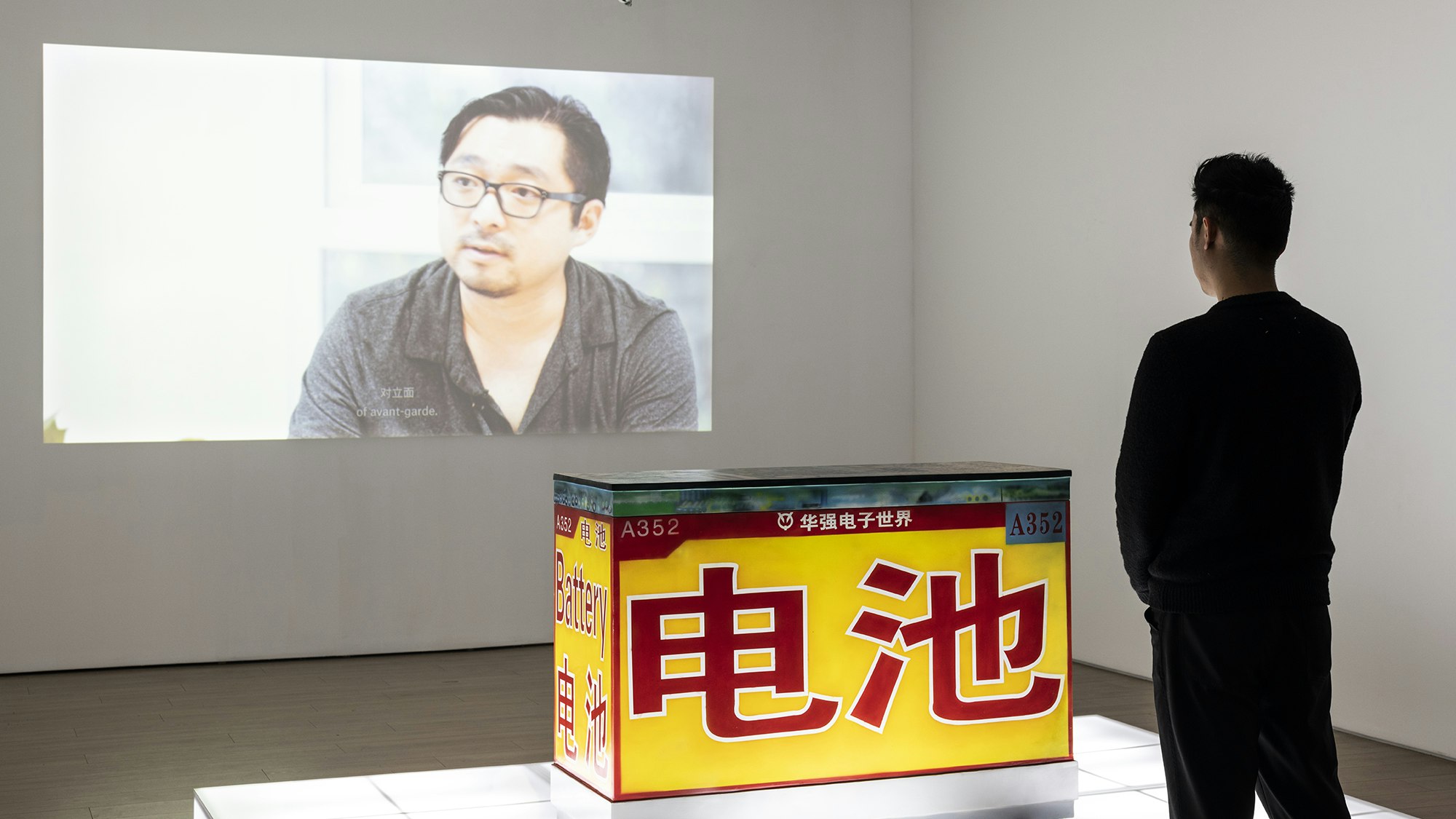 A male-presenting figure dressed in black stands in front of a yellow sculptural block airbrushed with the red Chinese character for 'Battery', situated on an illuminated white plinth. On the wall is a projection of a video still showing an East Asian male-presenting figure wearing glasses.