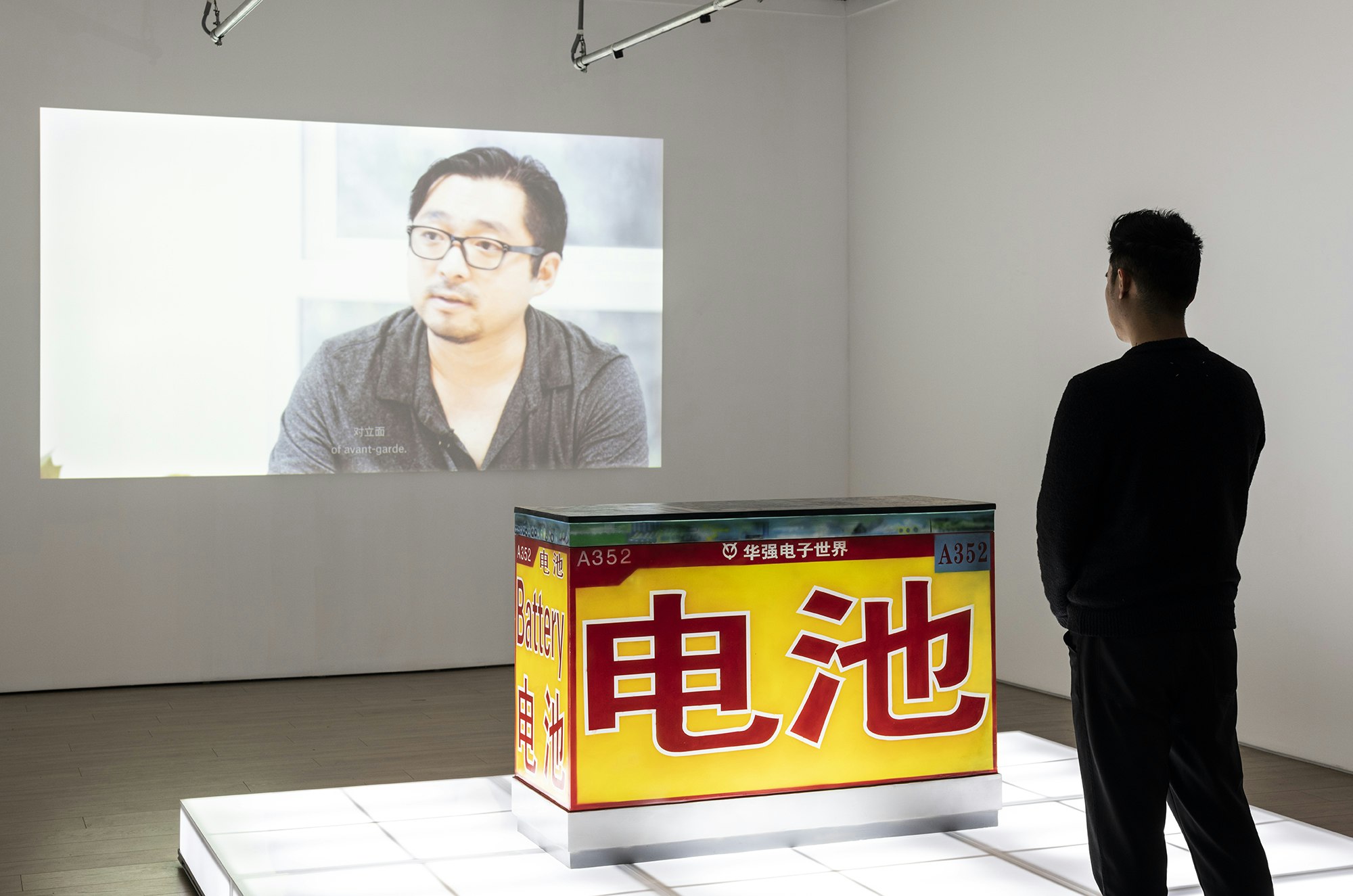 A male-presenting figure dressed in black stands in front of a yellow sculptural block airbrushed with the red Chinese character for 'Battery', situated on an illuminated white plinth. On the wall is a projection of a video still showing an East Asian male-presenting figure wearing glasses.