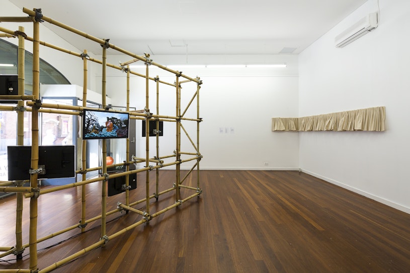A bamboo scaffolding structure mounted with television screens faces two white gallery walls hung with a short curtain of beige fabric
