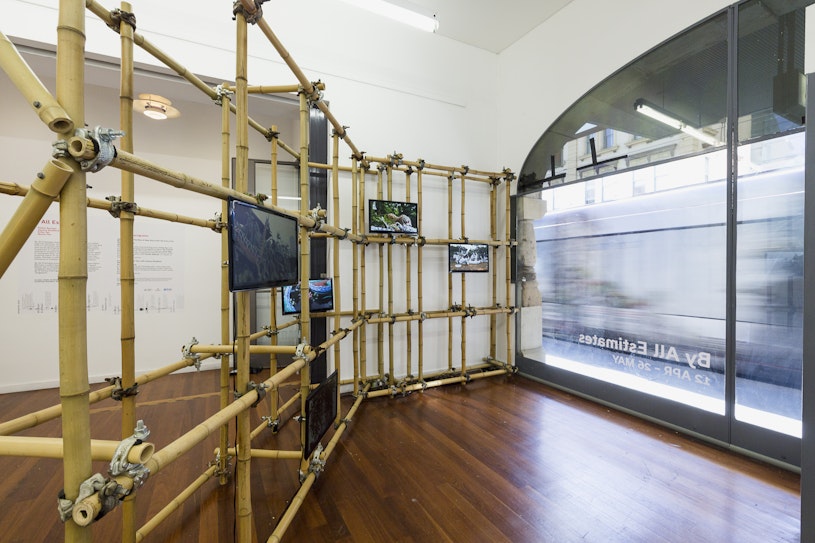A bamboo scaffolding structure mounted with five television screens faces a gallery window as a tram goes past outside