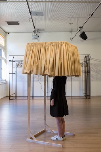 A figure in a black dress and white sneakers standing with their head stuck inside a wooden structure lined with a short beige curtain. Behind them is a colonial shopfront welded from steel and brass