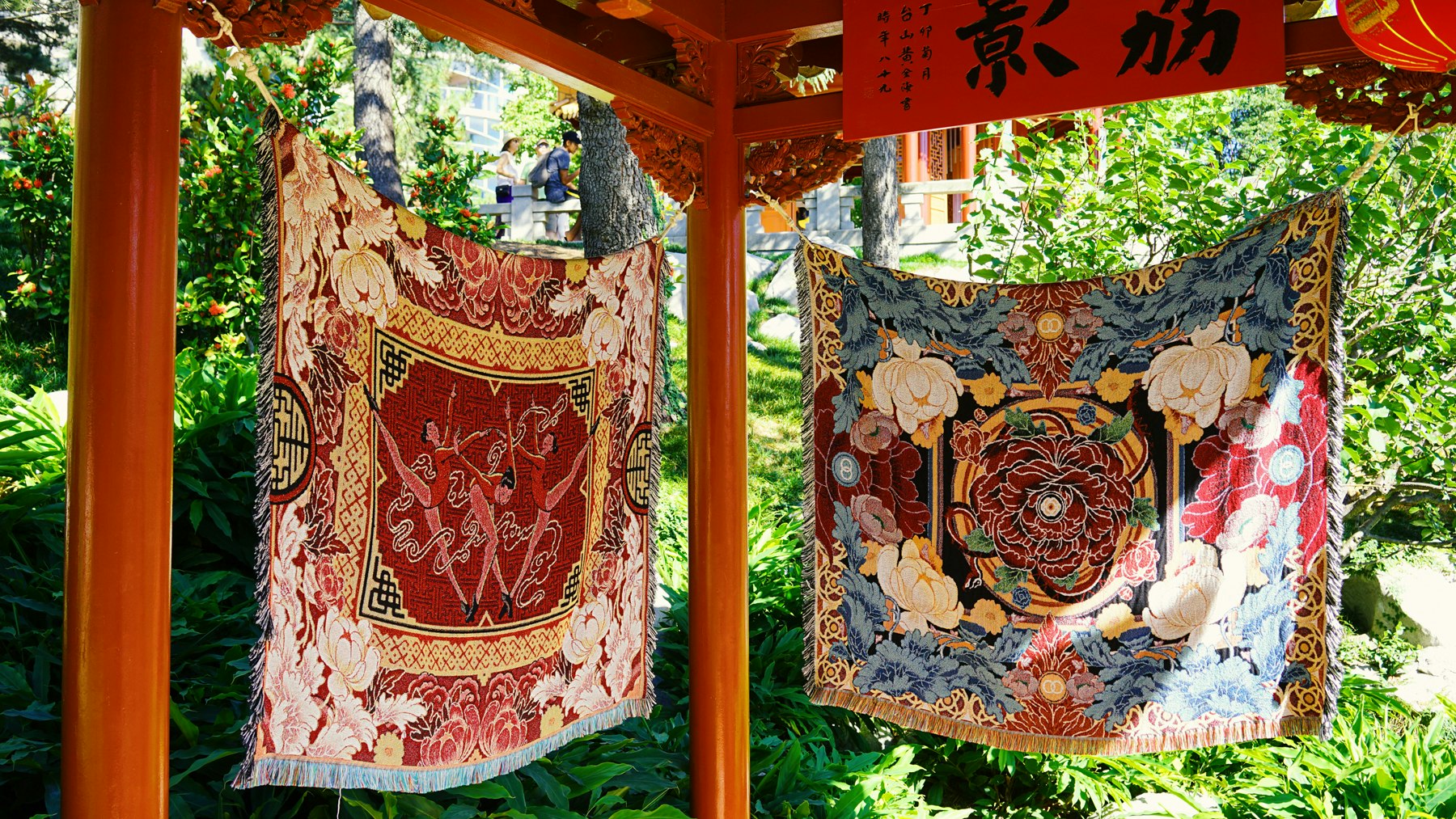 Two woven tapestries suspended from columns in a garden. The one of the left is woven from red, gold and pink colours with three dancers centred and framed by flowers. The one on the right is composed from red and white petals, and blue leaves.