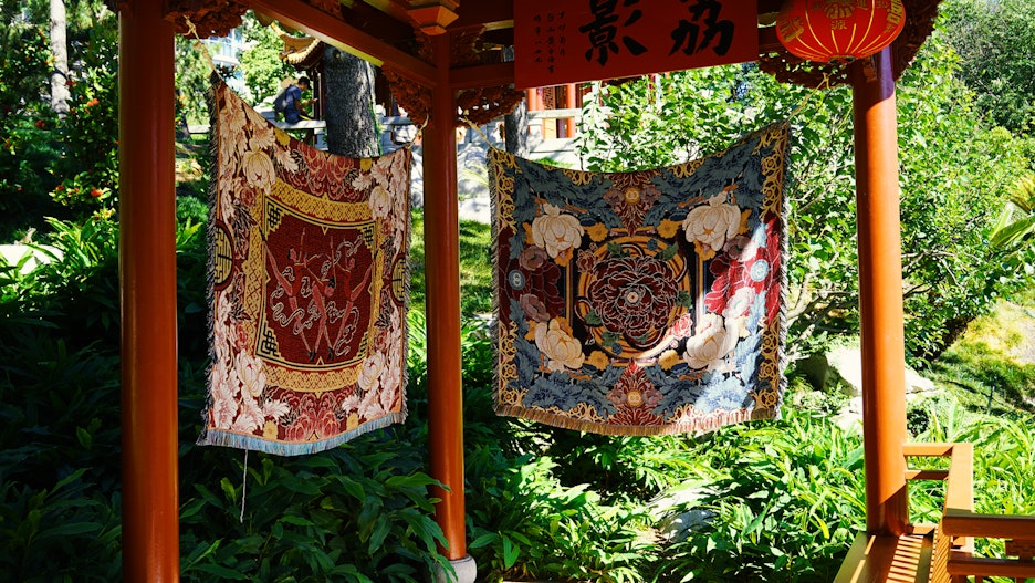 Two woven tapestries suspended from columns in a garden. The one of the left is woven from red, gold and pink colours with three dancers centred and framed by flowers. The one on the right is composed from red and white petals, and blue leaves.