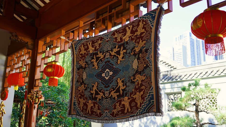 A woven tapestry of children dancing, suspended next to red lanterns in a garden