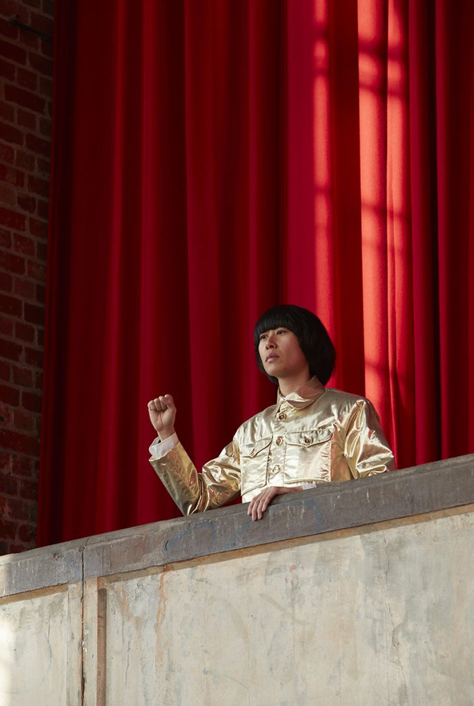 A female-presenting figure stands in a gold uniform, leaning over a concrete balcony. She holds up a closed fist. Behind her is a tall red curtain.