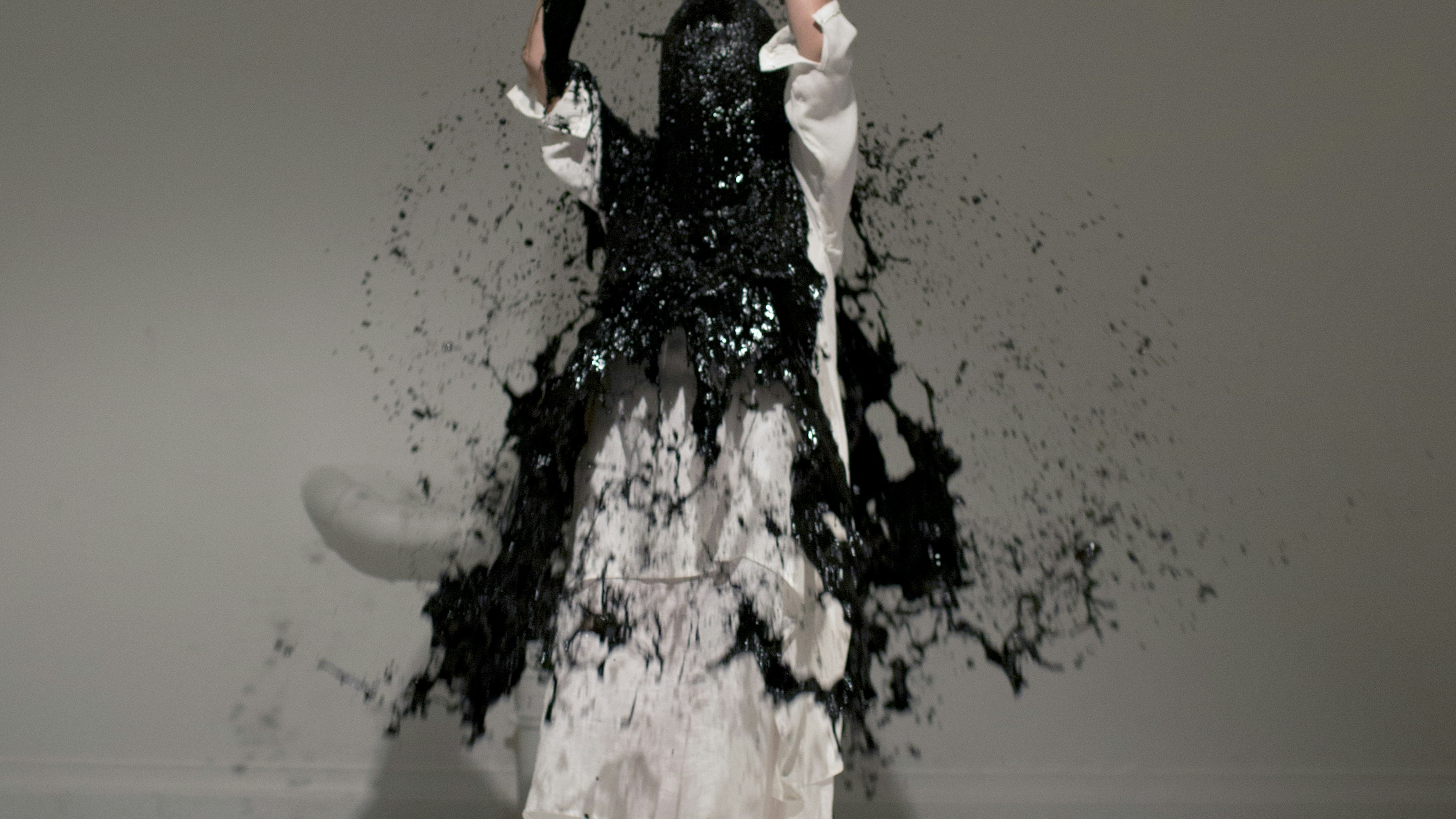 A photo of artist Xiao Lu, standing in a white dress raising a black bucket above her head and black paint splashing across her head and body.