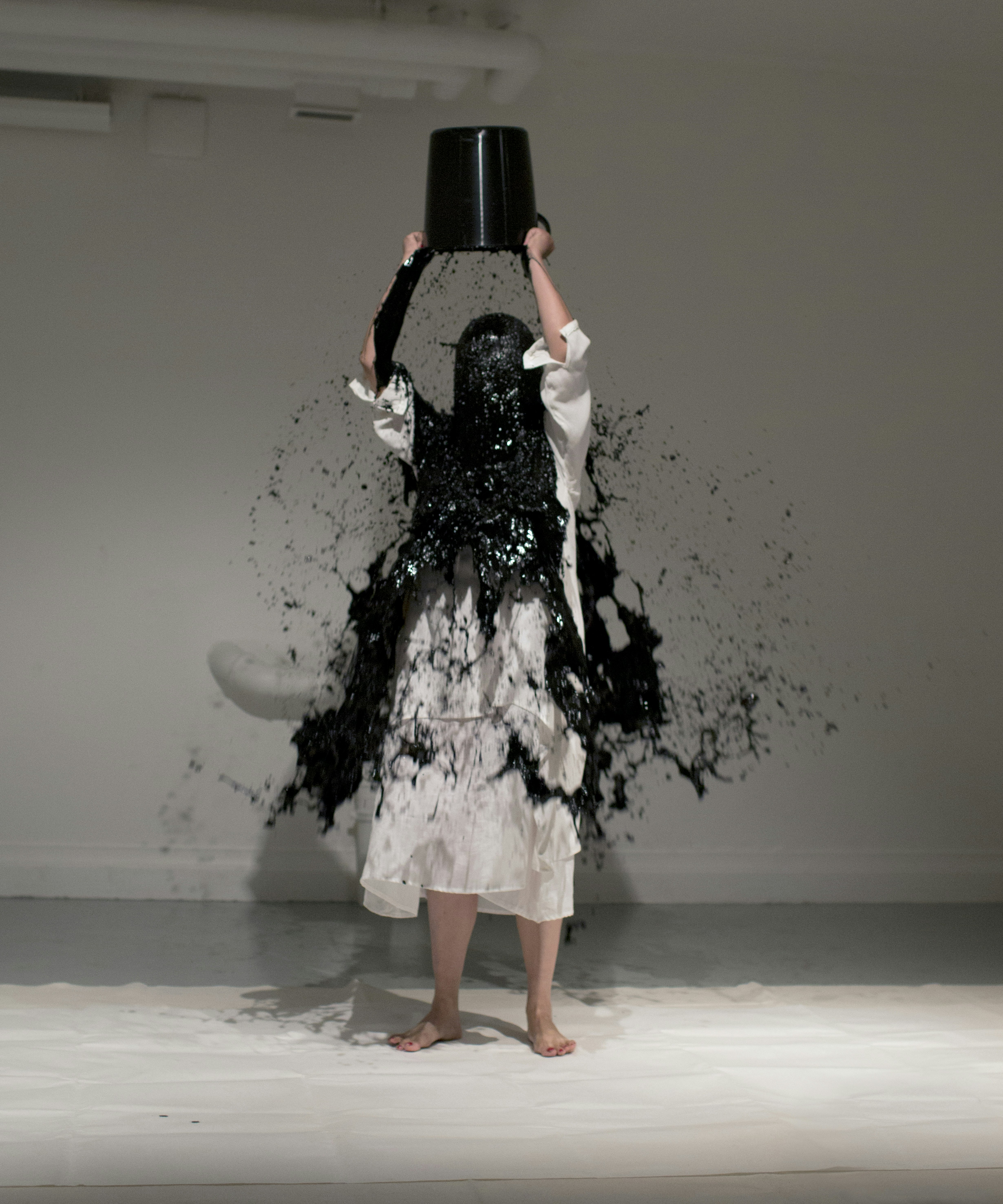 A photo of artist Xiao Lu, standing in a white dress raising a black bucket above her head and black paint splashing across her head and body.