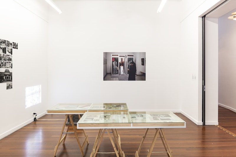 Two glass display cabinets in a white gallery space, with a photographic print of a woman in black with a ponytail, shooting a pistol. On the left wall is a projection and a collection of black inkjet prints