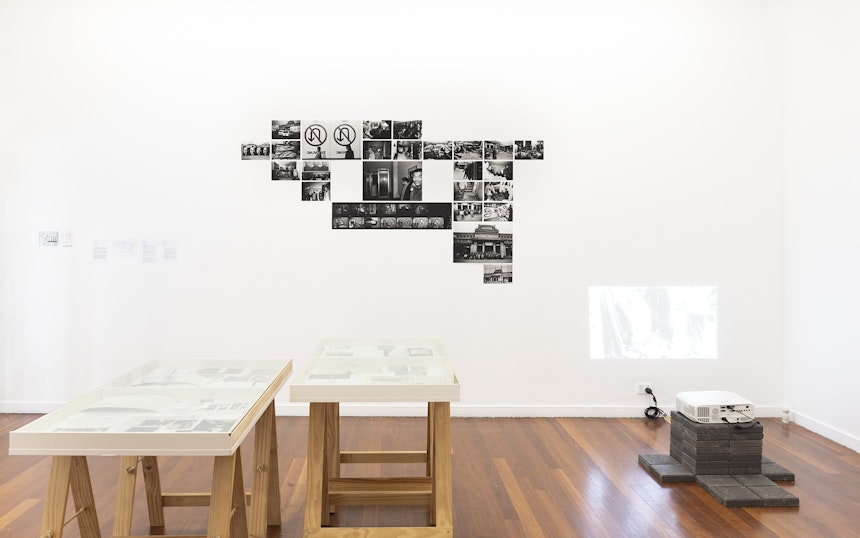 A white gallery space with two glass display cabinets, a projector stacked on concrete blocks facing a wall and a selection of black inkjet photographs pasted up on the wall