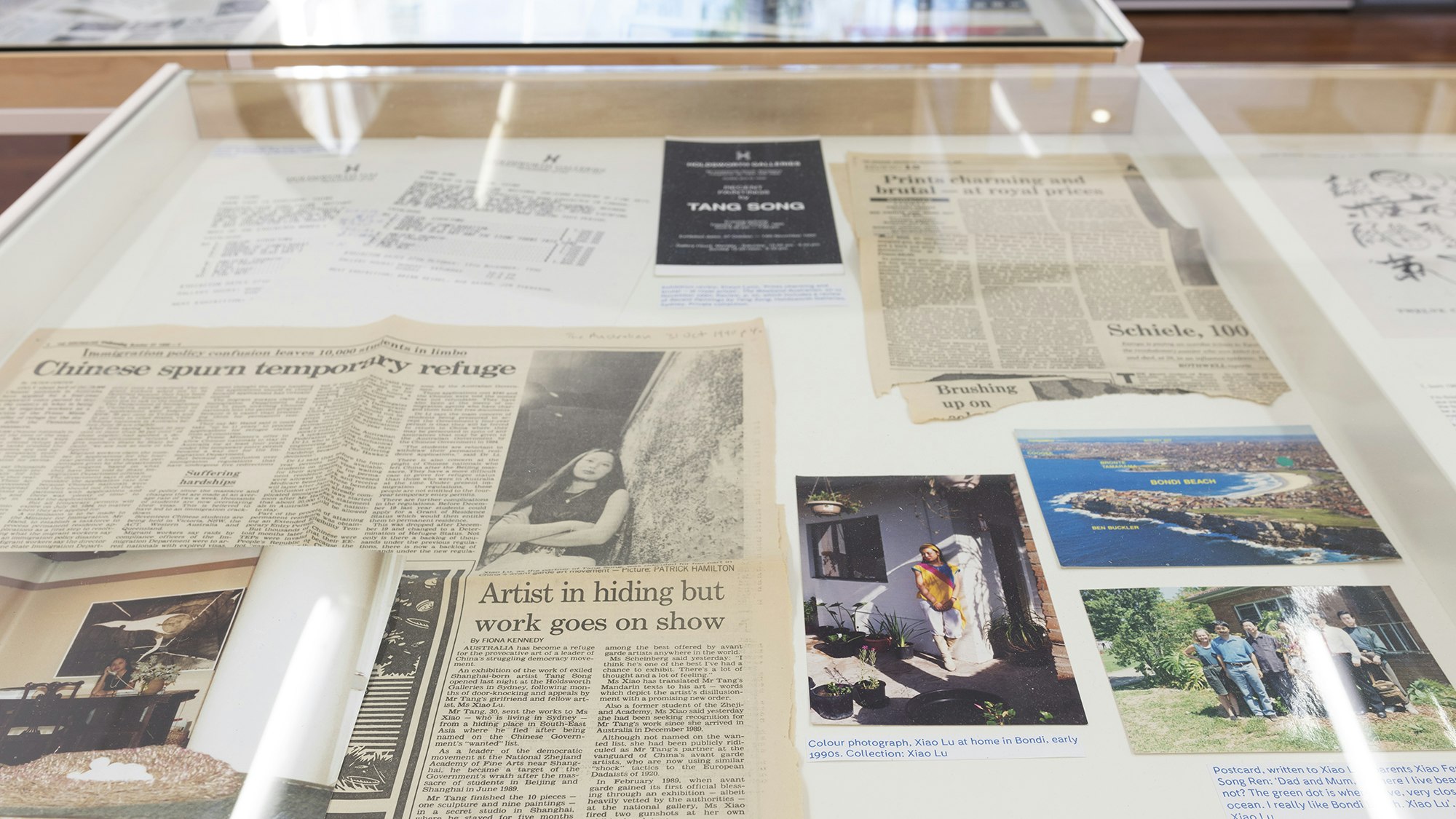 Yellowed newspaper clippings and photo prints in a glass display cabinet, one of the clippings titled 'Artist in hiding but work goes on show'