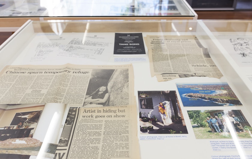 Yellowed newspaper clippings and photo prints in a glass display cabinet, one of the clippings titled 'Artist in hiding but work goes on show'