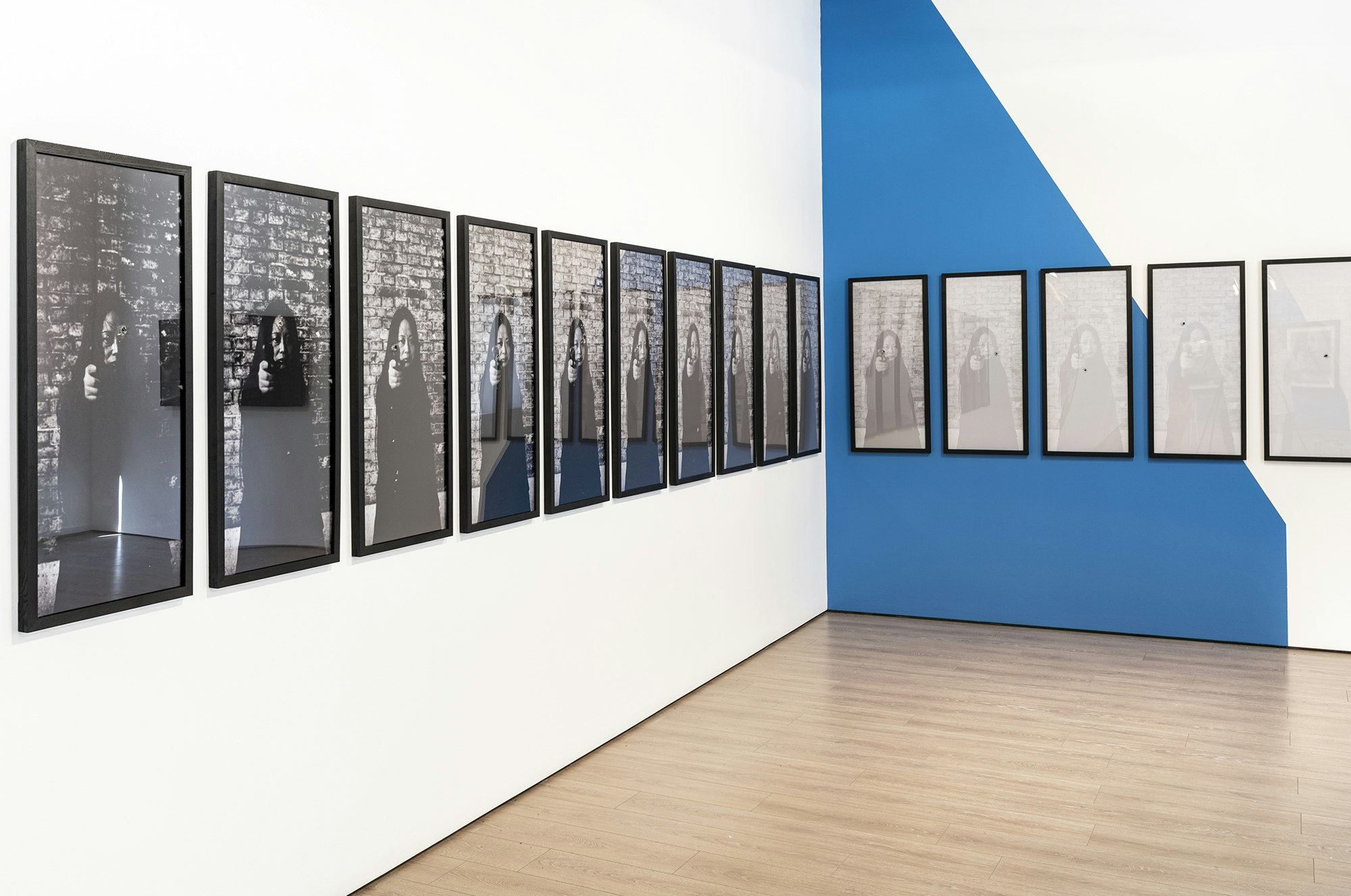 Fifteen framed black and white prints depicting an East Asian woman with long black hair, torso up, standing against a brick wall and pointing a pistol at the camera