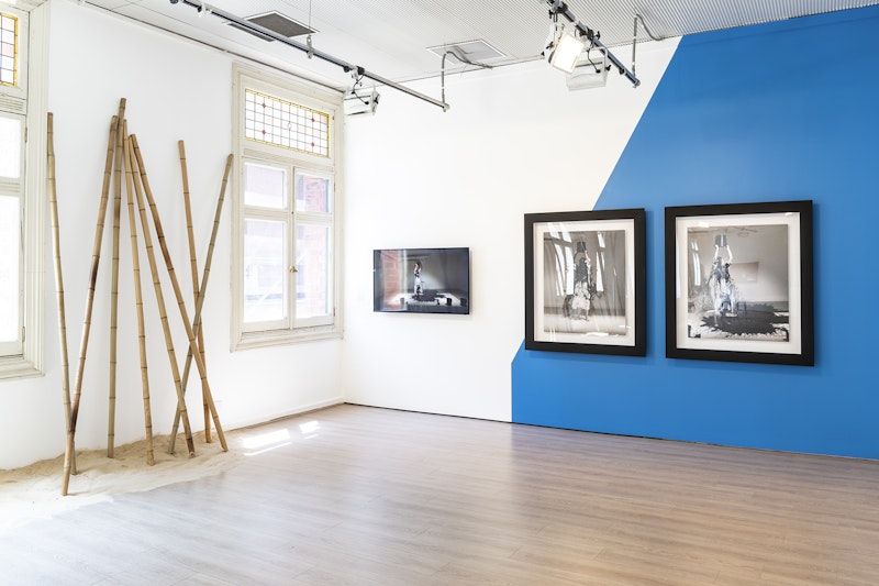 A corner of a white gallery space, with part of the wall painted blue, showing two large colour prints of a figure in a white dress tipping a bucket of black paint over her head and her body. A video screen next to the prints shows a video recording of a woman in a white dress pouring a bucket of black paint over her head. On the left wall are several bamboo poles stood upright