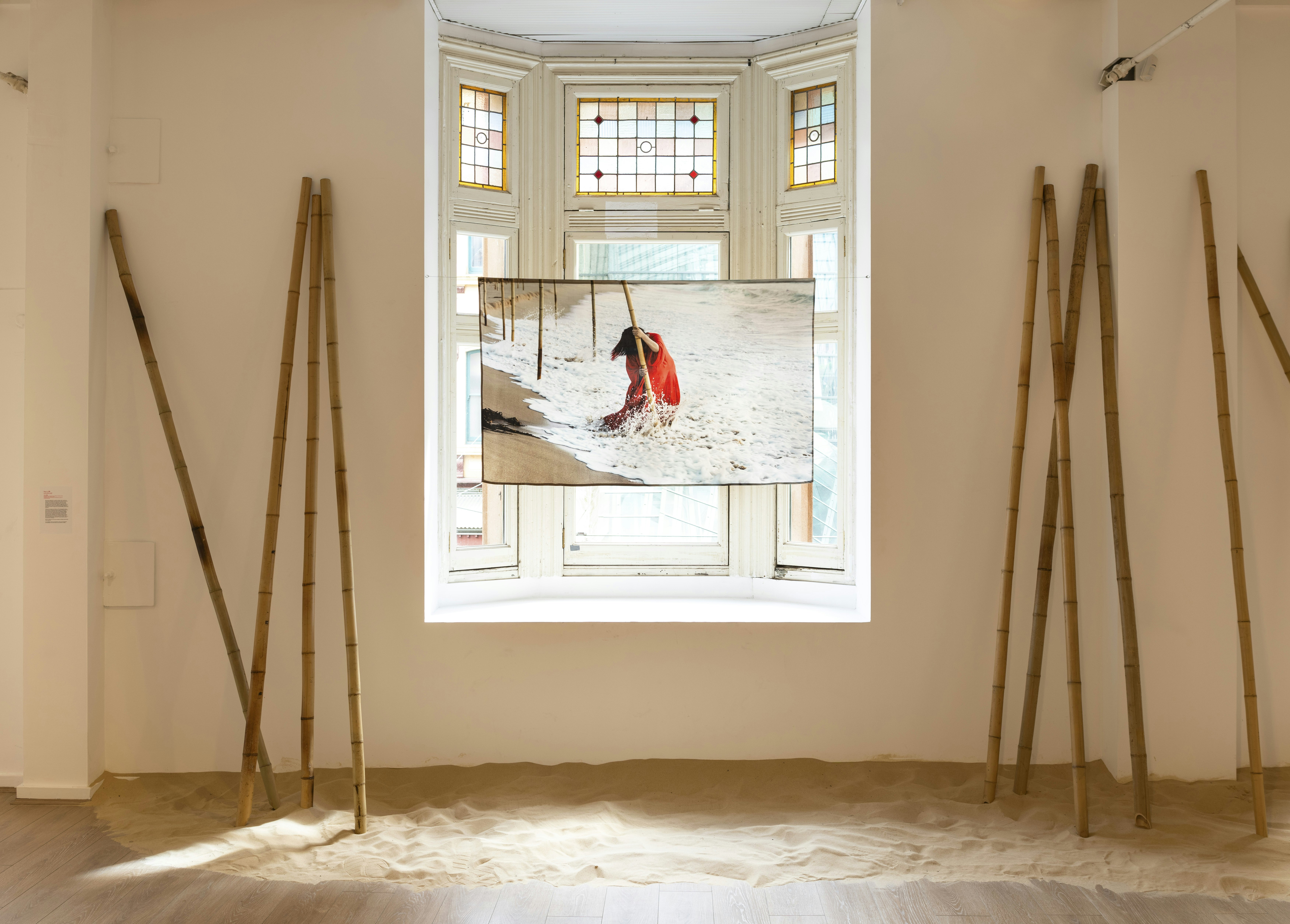 A video screen showing a female-presenting figure in a red dress standing knee-deep in the ocean grasping a bamboo pole in both of her hands. The screen is suspended in front of some stained glass windows, with several bamboo poles standing upright on each side of the windows.