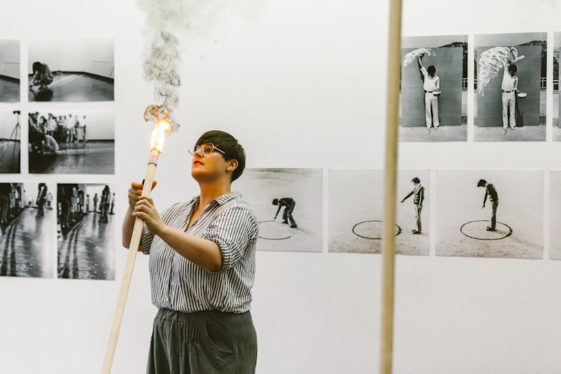 Emily, a femme-presenting figure with short cropped hair and glasses sets fire to a wooden pole. Behind her are black and white prints of a man drawing a circle on the ground and scribbling on a wall.